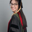 Young woman with glasses wearing doctorate regalia turns and smiles at the camera. She has long black hair and is standing in front of a gray background.